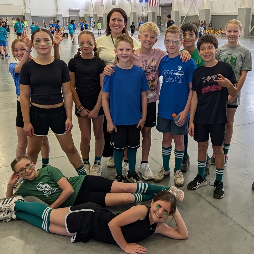 Bountiful Elementary students and a staff member pose together for a team photo at the district Decathlon. Students wear athletic clothes, school colors, striped socks, and face paint while standing and sitting together inside the large competition space.