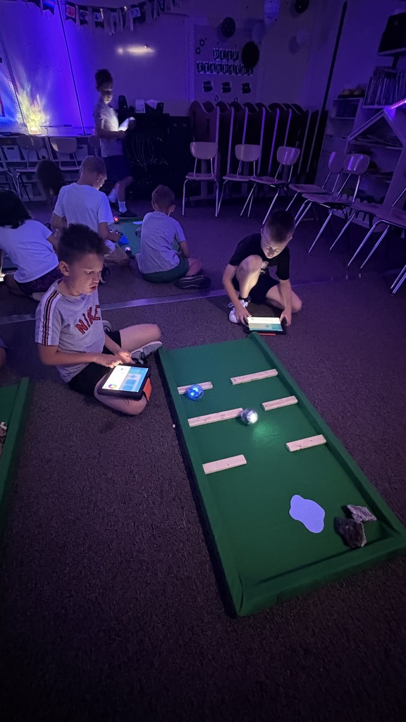 two boys sitting on the ground using ipads to control robots on a golf course