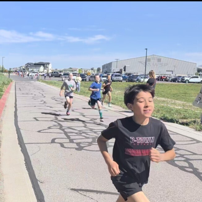 A Bountiful Elementary student runs outside during the district Decathlon, with teammates following behind on the course. The students are participating in an outdoor running event near the athletic facility on a sunny day.