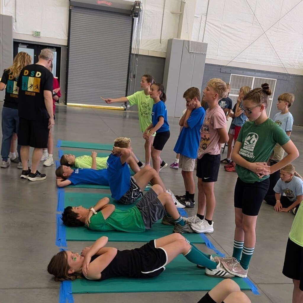 Bountiful Elementary students participate in the curl-up event during the district Decathlon. Several students lie on exercise mats while teammates and event helpers stand nearby, supporting and observing the activity.