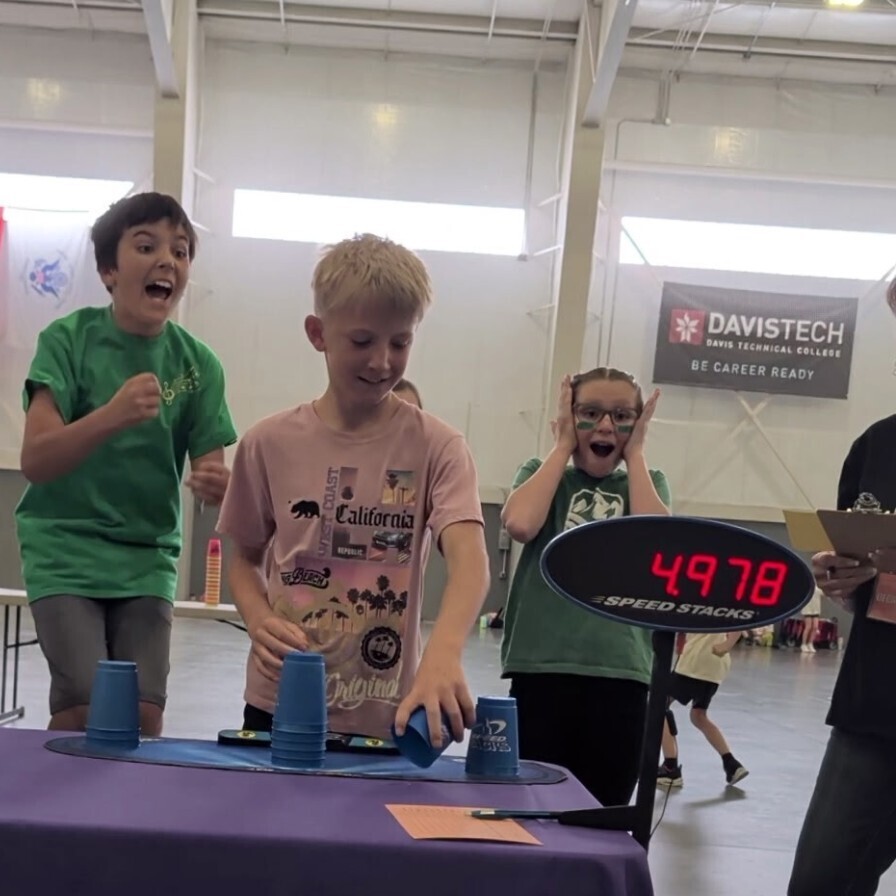 A Bountiful Elementary student participates in the cup stacking event at the district Decathlon while teammates react with excitement nearby. A digital Speed Stacks timer is visible beside the table, showing a completed time.