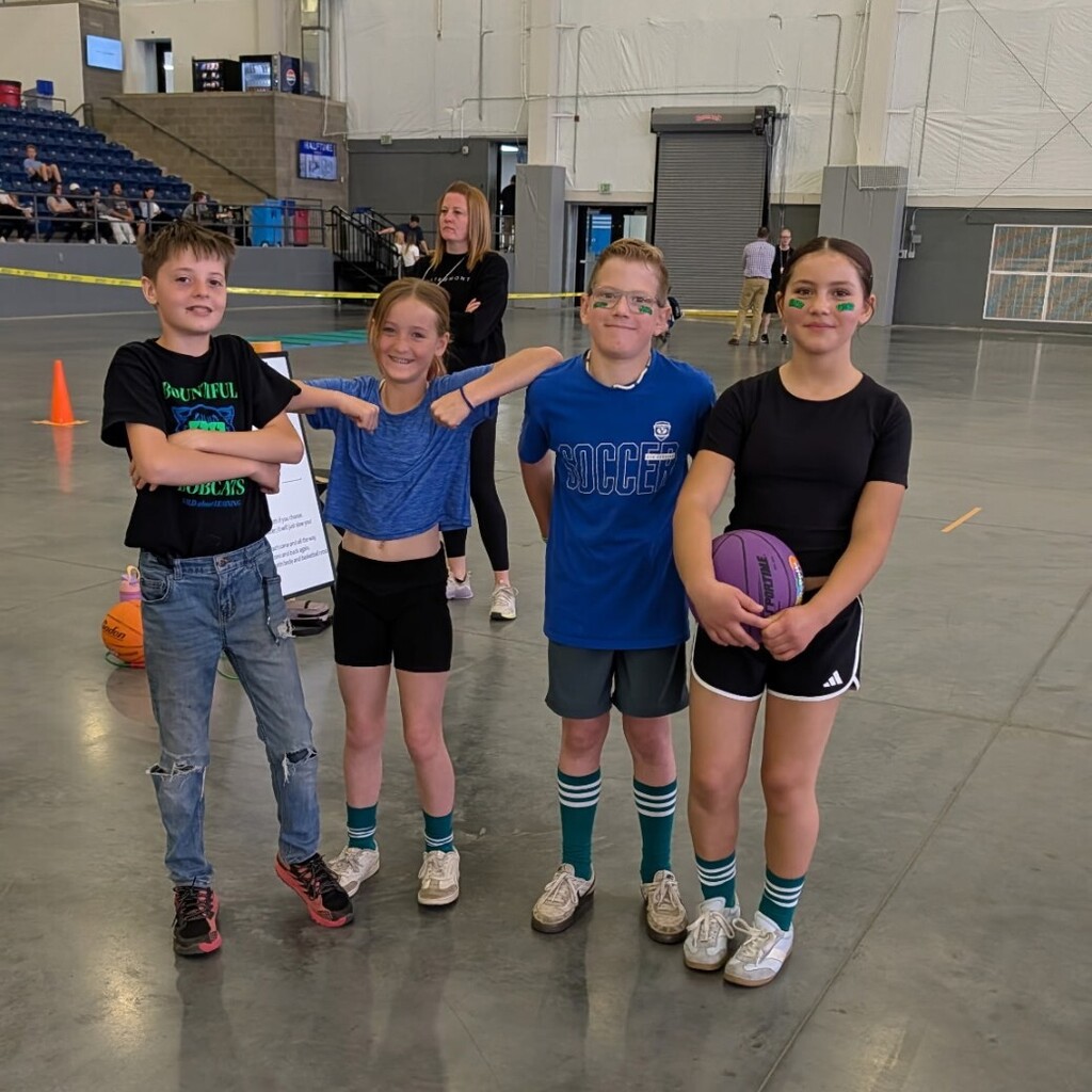Four Bountiful Elementary students pose together at the district Decathlon event inside a large indoor athletic facility.