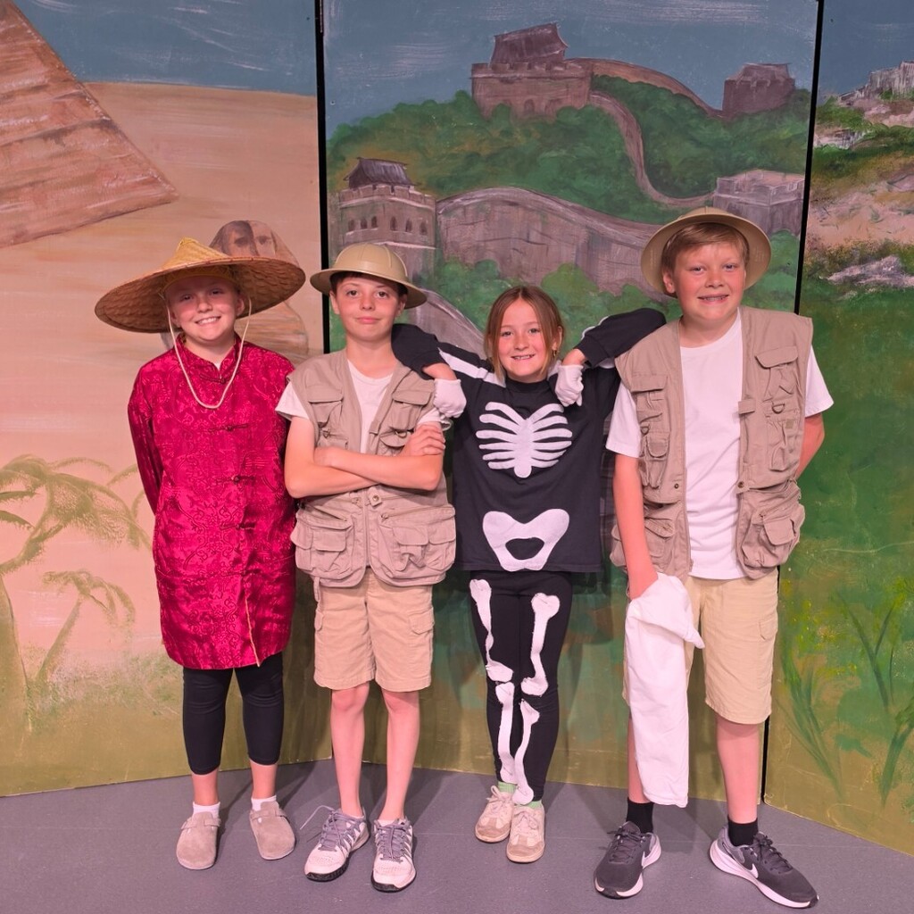 Four Bountiful Elementary 6th grade students pose on stage in costumes representing explorers and ancient civilizations. The group includes students wearing a red robe, safari-style vests and hats, and a skeleton costume in front of the “Dig It” performance backdrop.