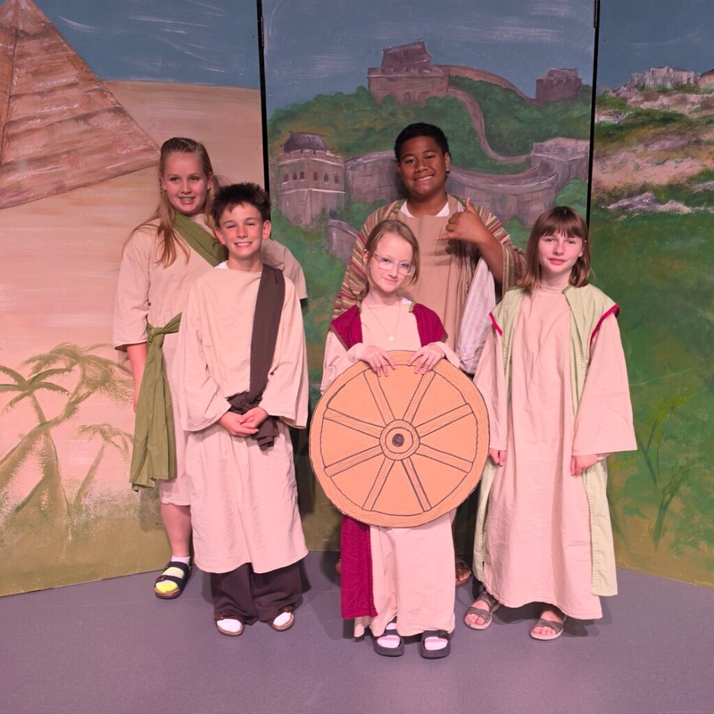 Five Bountiful Elementary 6th grade students pose on stage in ancient civilization costumes. One student holds a large cardboard wheel prop, while others wear robe-style costumes in front of a painted backdrop featuring a pyramid, the Great Wall of China, and ancient ruins.
