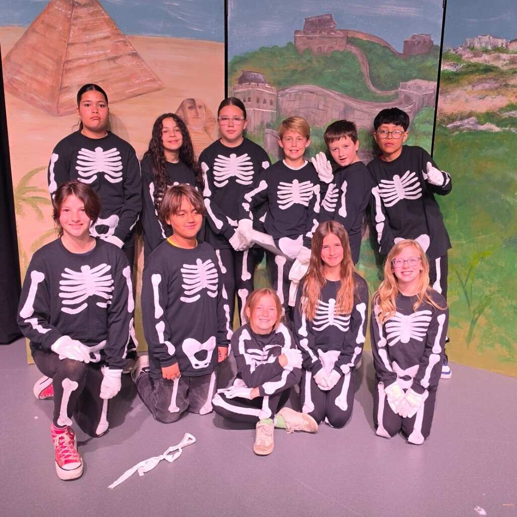A group of Bountiful Elementary 6th grade students pose together on stage wearing matching black skeleton costumes with white bone designs. They are in front of the painted ancient civilizations backdrop used for the 6th grade “Dig It” performance.