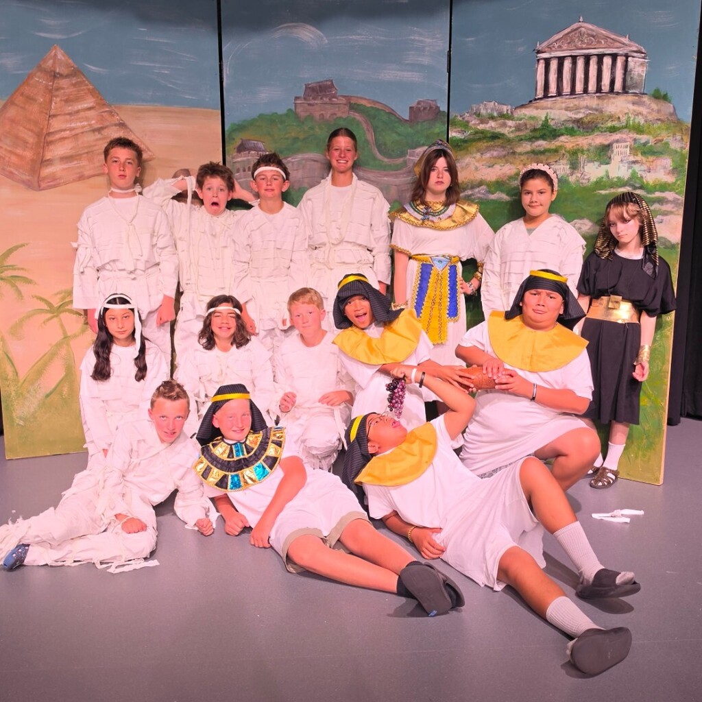group of Bountiful Elementary 6th grade students pose on stage in costumes inspired by ancient Egypt and mummies. Some students wear white wrapped costumes, while others wear Egyptian-style collars, headdresses, and robes in front of the “Dig It” ancient civilizations backdrop.