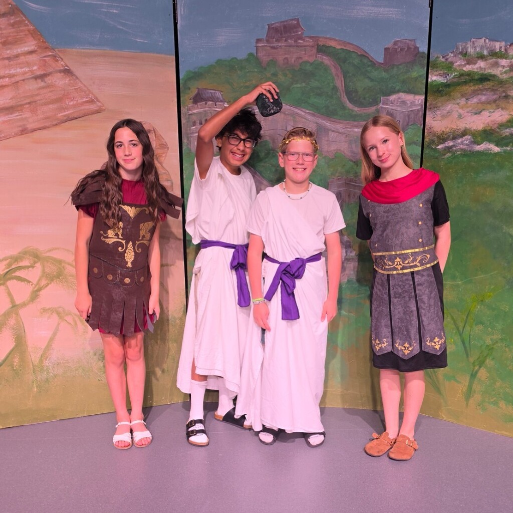 Four Bountiful Elementary 6th grade students pose on stage in costumes for the “Dig It” performance. Two students wear Roman-style armor costumes, while two others wear white Greek-style costumes with purple sashes in front of a painted ancient civilizations backdrop.