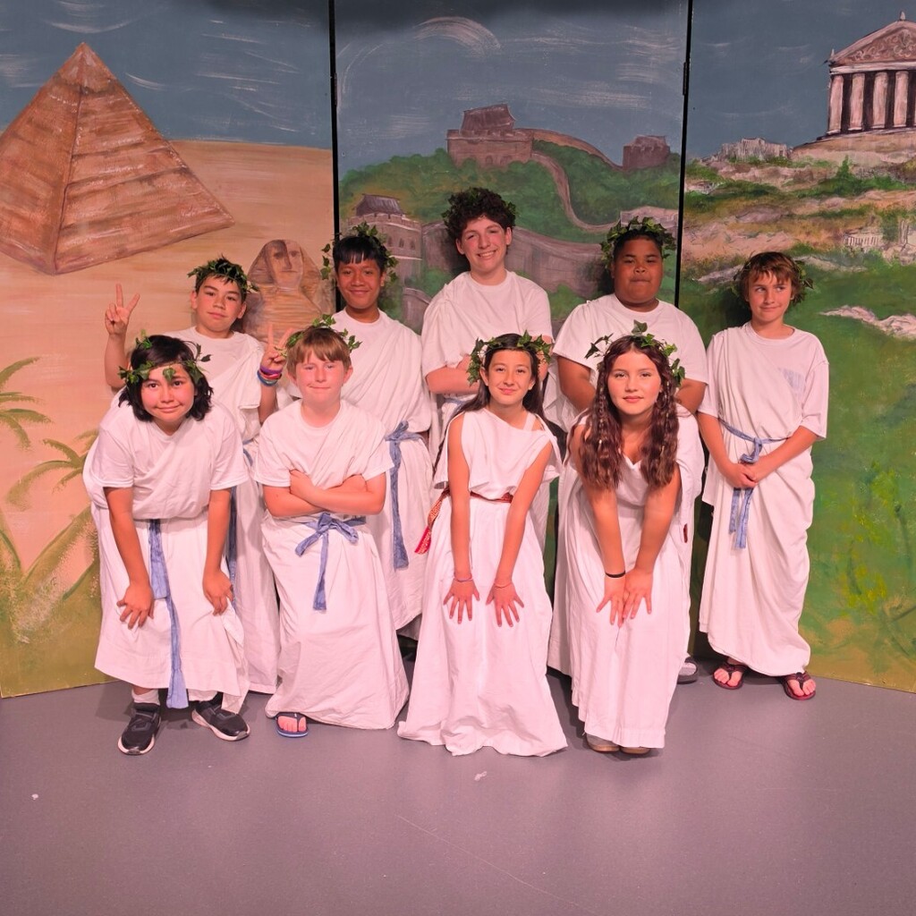 Bountiful Elementary 6th grade students pose on stage in white Greek-style costumes with leafy headpieces. Behind them is a painted backdrop showing ancient landmarks, including a pyramid, the Great Wall of China, and a Greek temple, for their “Dig It” performance about ancient civilizations.