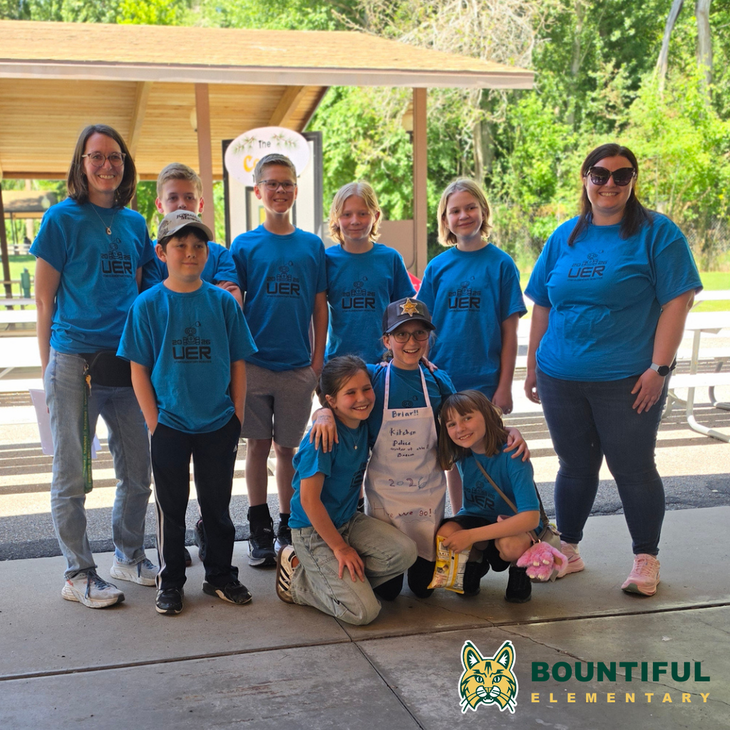 Bountiful Elementary UER Robotics students and coaches pose together at Lagoon during USU’s Physics Day. The group wears matching blue UER shirts and stands under a pavilion with trees and picnic tables in the background.