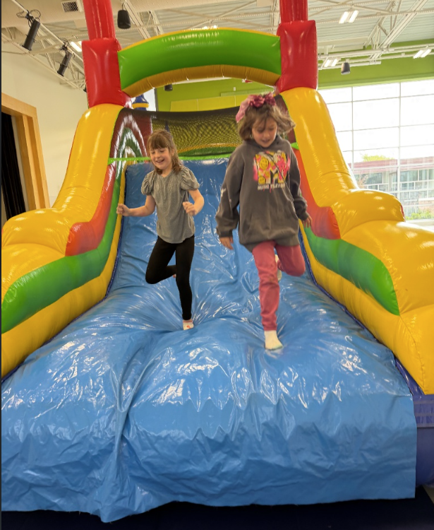 Two elementary school girls smile and laugh as they slide down a large, colorful indoor inflatable slide. The slide has bright yellow, red, and green walls with a wide blue sliding surface.