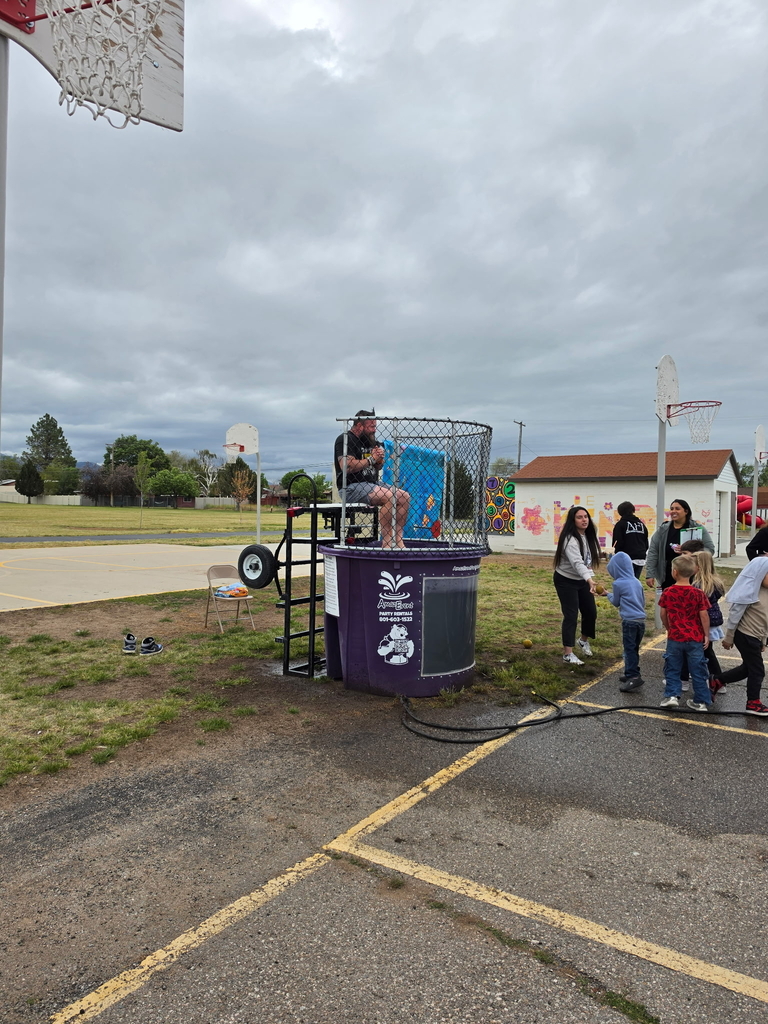 A man sits above a purple dunk tank on an asphalt playground under an overcast sky. A few young elementary school students stand in line waiting to throw a ball, while two adults look on. Basketball hoops and a grassy field are visible in the background.
