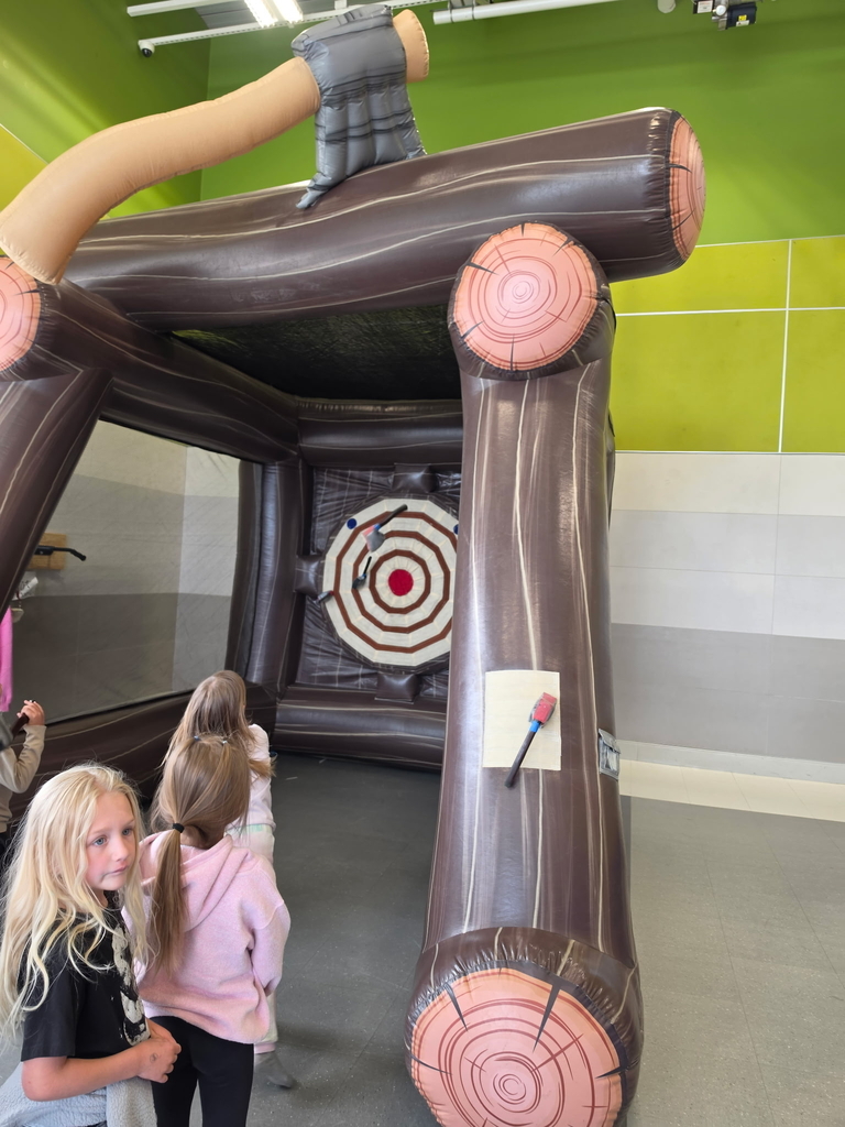 Two young girls watch an indoor inflatable axe throwing game. The large inflatable structure looks like a log cabin frame with a bullseye target at the back, where two foam axes are stuck to the hook-and-loop surface.