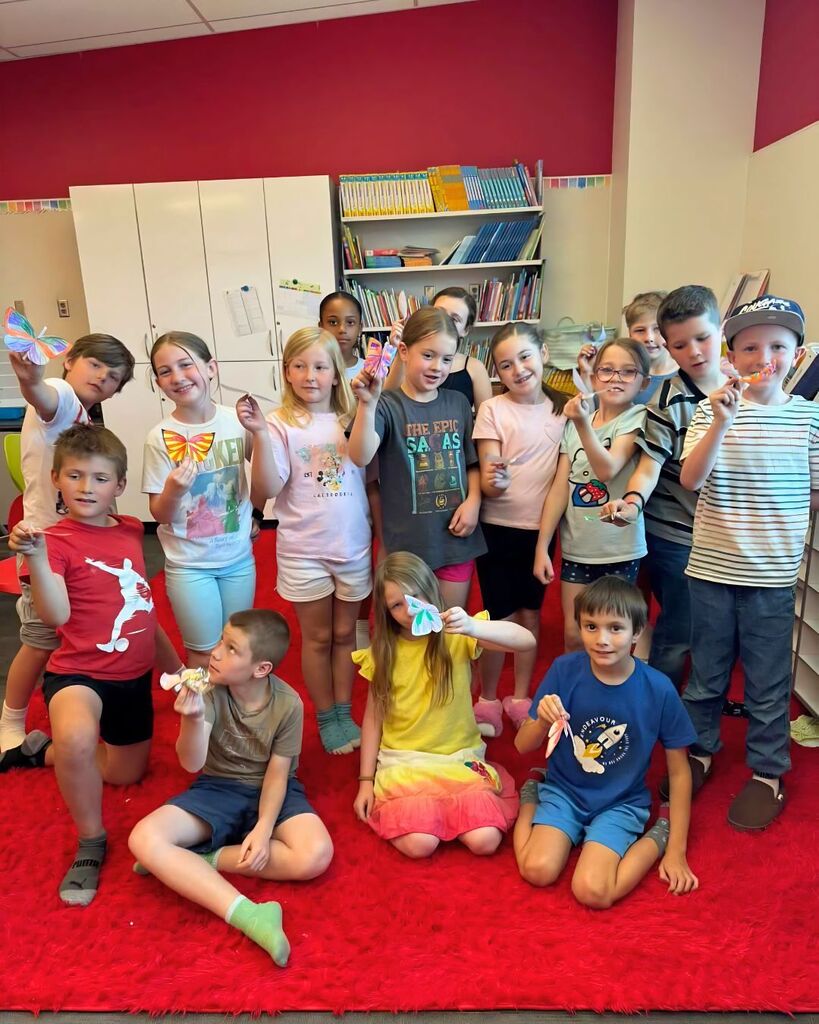 A group of elementary students pose together on a bright red classroom rug while holding colorful butterfly crafts they created. Some students kneel in the front while others stand behind them, smiling and showing off their artwork in a classroom filled with bookshelves and cabinets.