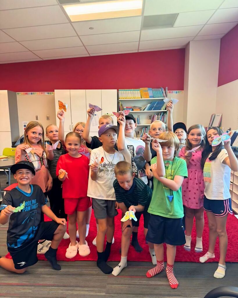 A group of elementary students pose together on a bright red classroom rug while holding colorful butterfly crafts they created. Some students kneel in the front while others stand behind them, smiling and showing off their artwork in a classroom filled with bookshelves and cabinets.