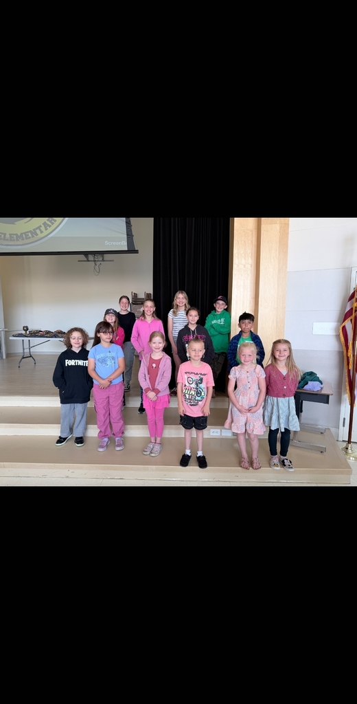 A group of eleven elementary school students stand in two neat rows on a wooden stage during a school assembly. A projector screen is lowered behind them on the left, and a dark curtain hangs on the right.