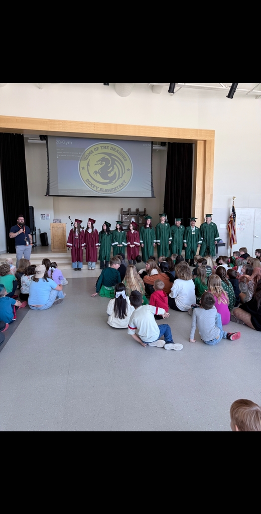 A group of ten high school seniors wearing green and maroon graduation caps and gowns stand in a line on the school stage. A male faculty member speaks into a microphone on the left. A large audience of elementary school students sits on the gymnasium floor watching them. The projector screen above the stage displays the Doxey Elementary Dragons logo.