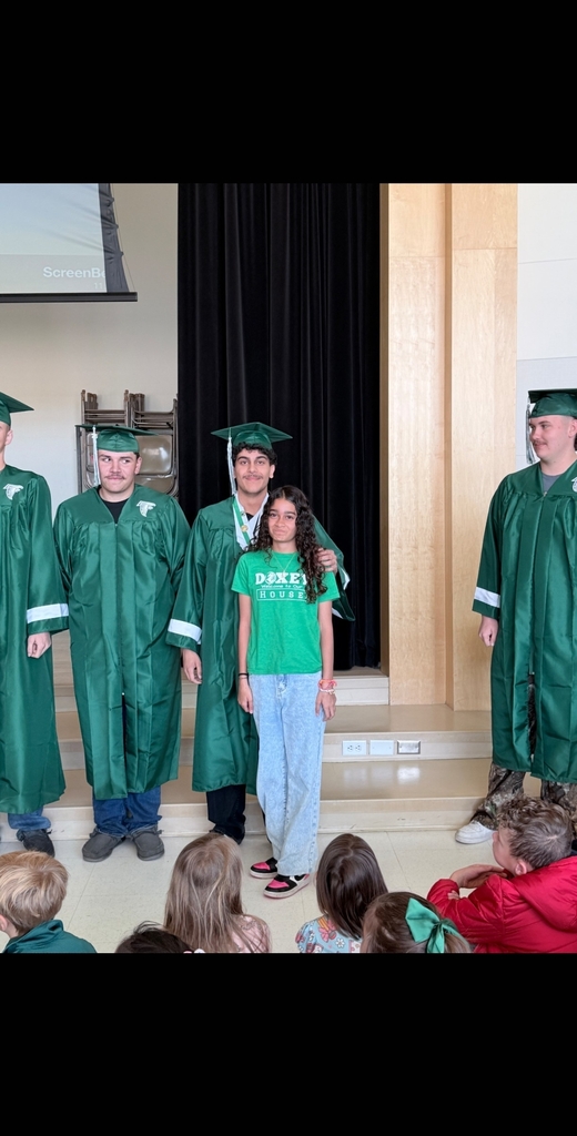 A close-up shot of four high school seniors in green graduation gowns and caps standing on the stage. A younger elementary school girl in a green Doxey shirt stands proudly in front of one of the graduates, who has a supportive hand on her shoulder.