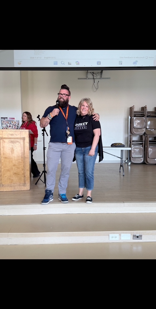 A bearded male faculty member wearing glasses and an orange lanyard stands on stage speaking into a microphone. He has his arm around Sherry Tweed, who is smiling warmly and wearing a black Doxey shirt.