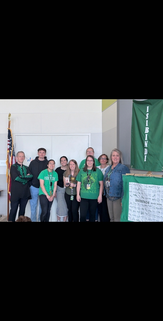A group of nine school faculty and staff members stand together on the stage celebrating. Several members are wearing green "Isibindi" shirts. To their right, a green house banner hanging on the wall reads "ISIBINDI" vertically, above a signed white banner.
