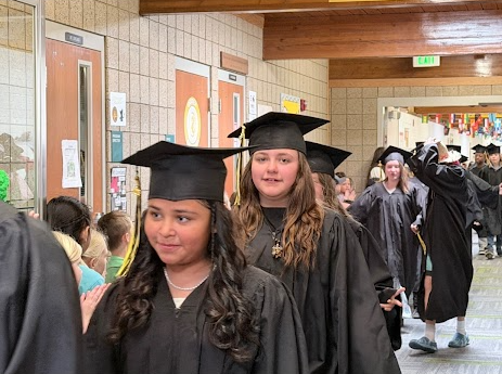 A profile view of a student in a black graduation gown and cap walking down the school hallway, looking toward cheering onlookers. Other graduates in gowns follow closely behind them.