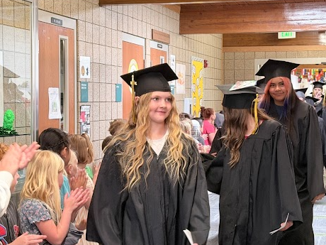 A smiling graduate with long blonde hair walks down the school hallway in a cap and gown. On the left, younger elementary students stand along the wall, smiling and clapping for the passing parade.