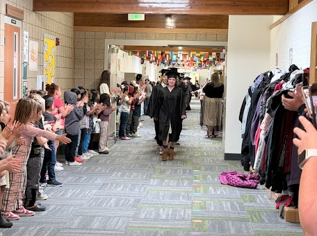 A wide shot down a long elementary school hallway lined with international flags hanging from the ceiling. Younger students line both sides of the corridor, cheering and high-fiving a procession of graduates walking down the center in caps and gowns.