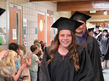 A bright, smiling graduate with long brown hair walks down the hallway in a black cap and gown, while younger students line the wall on the left to offer high-fives and applause.