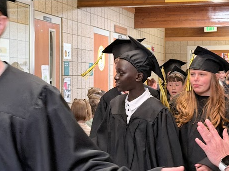 Two smiling elementary school graduates in black caps and gowns lead a line down a school corridor. Younger students line the left side of the hallway, watching and clapping as the graduates pass by.