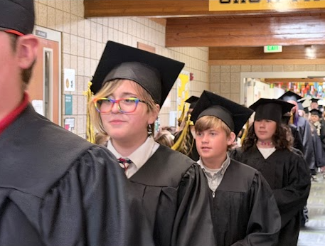A graduate wearing colorful rainbow-rimmed glasses, a white collared shirt, and a tie under their black cap and gown walks in a line down the hallway with a slight smile.