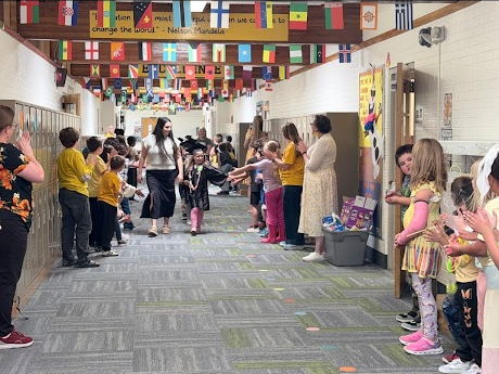 A wide perspective of the decorated school hallway where teachers and younger students stand along lockers and walls, clapping for a small graduate being escorted down the center of the hall by an adult. International flags hang across the wooden ceiling beams above.