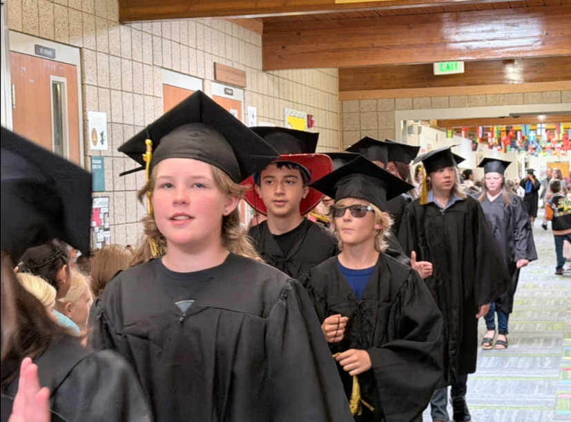 A close-up shot of elementary school graduates wearing black caps and gowns walking down a school hallway during a graduation parade. A student in the foreground looks ahead, while another behind them wears a red cowboy hat under their graduation cap, and a student next to them wears sunglasses.