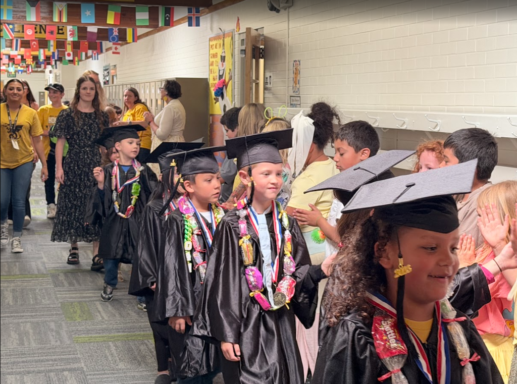 A view down a decorated school hallway where a line of kindergarten graduates in black caps and gowns walks down the center. Many of the children are wearing colorful candy leis and medals around their necks. Teachers and older students line the walls, clapping and cheering as the procession passes under international flags hanging from the ceiling.