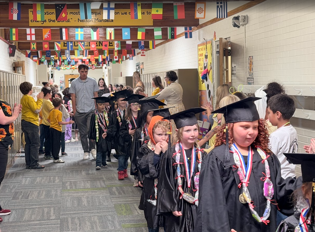 A line of kindergarten graduates wearing black graduation caps and gowns walks down an elementary school corridor. The students in front are wearing handmade candy leis over their gowns. On the sides of the hallway, other students and staff stand by the lockers, smiling and applauding the graduates.