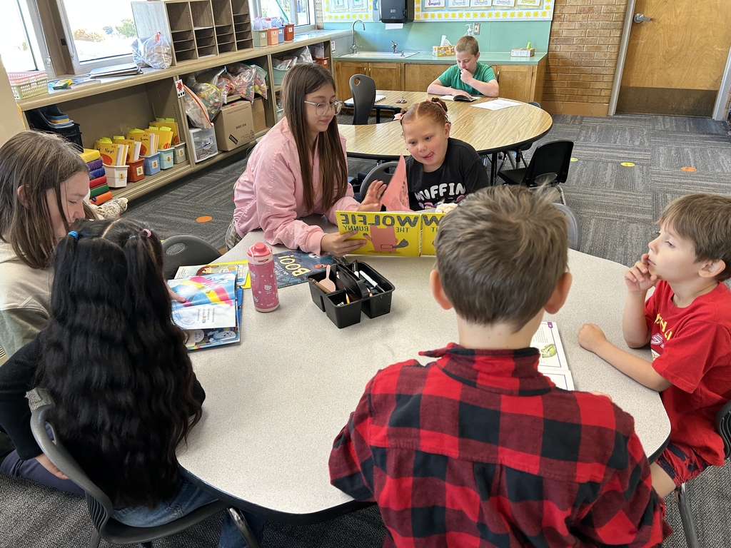 A group of elementary school students gathered around a large table inside a bright classroom, actively engaged in a reading activity. Several children are paired up or reading in small groups, including an older student in a pink jacket sharing a yellow children's book with a younger girl. Other students are independently looking through picture books or listening intently. The background shows classroom storage cubbies filled with books and learning materials, large windows, and a student working at another desk.