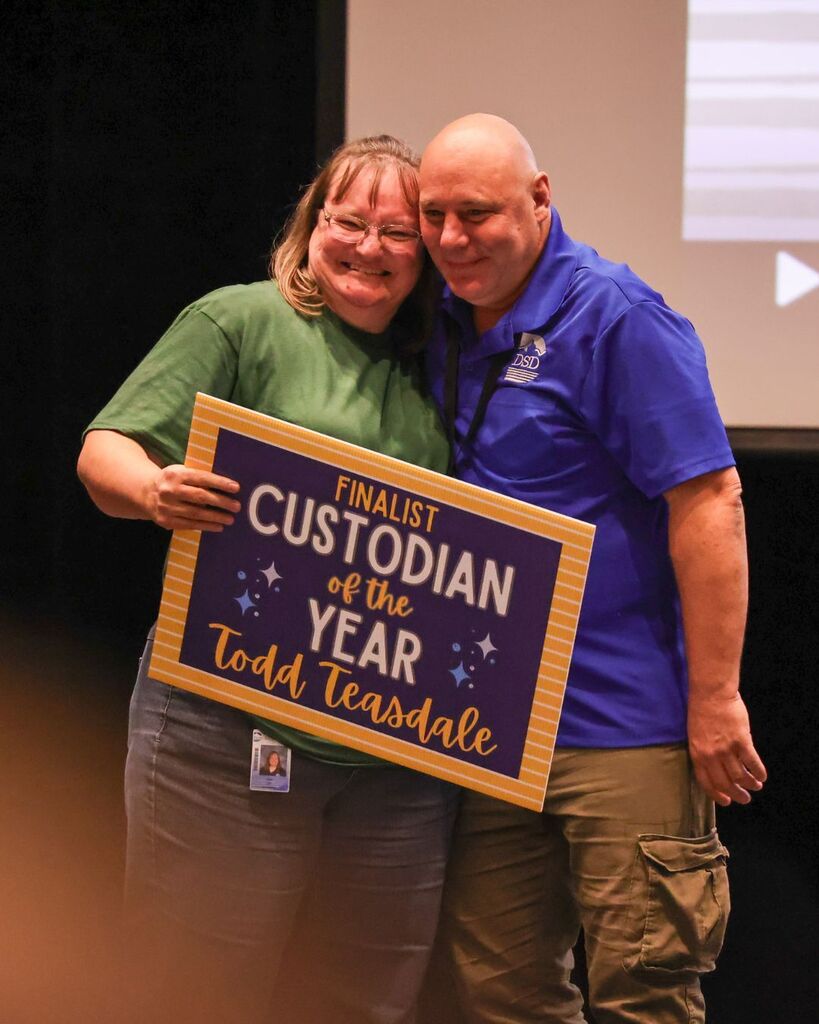 Custodians hugging. One is holding a sign that reads: "Finalist Custodian of the Year Todd Teasdale."