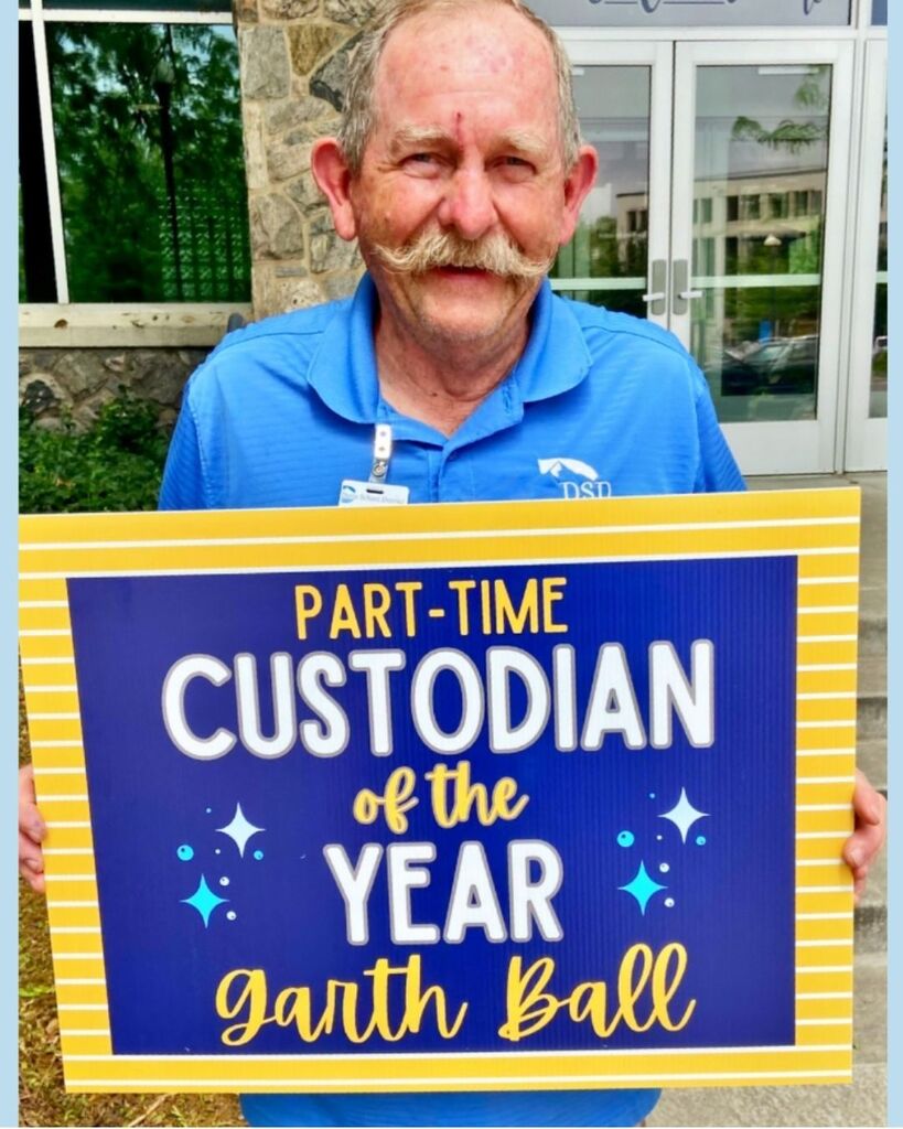 A DSD custodian holding up a sign that reads: "Part-time Custodian of the Year Garth Ball"