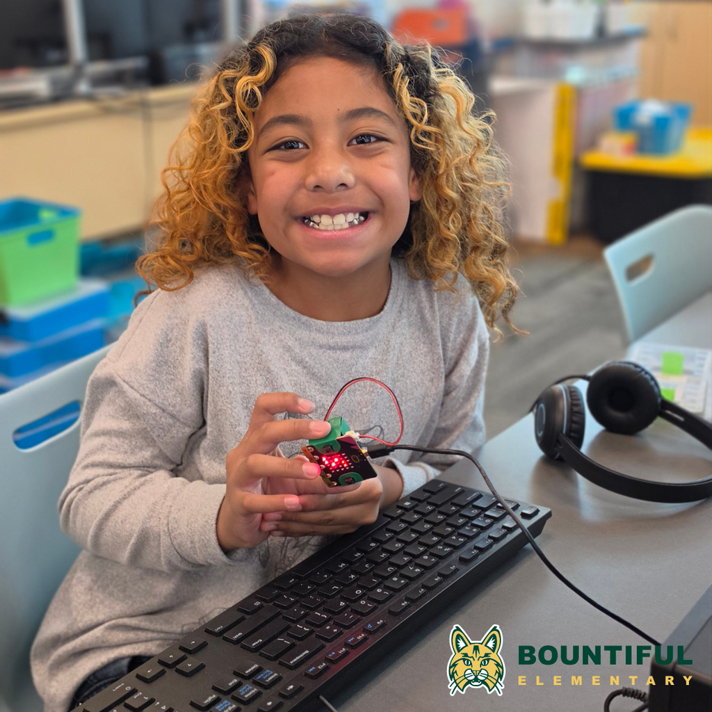 Bountiful elementary student smiling while holding a lit micro:bit connected to a computer during summer school.