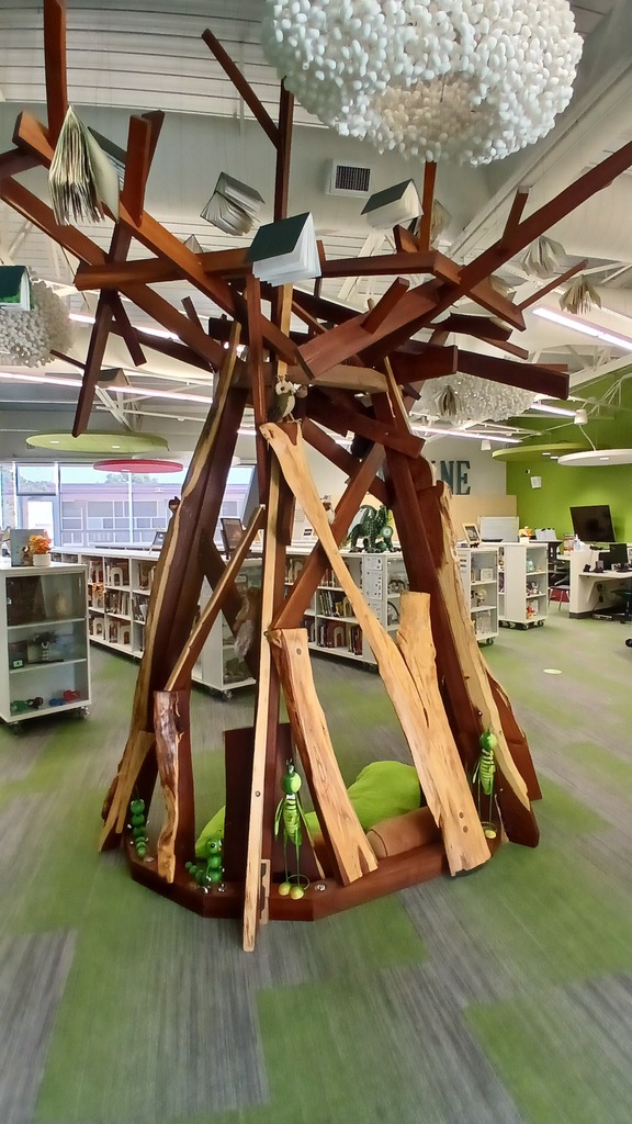 An indoor architectural wooden reading structure shaped like a stylized tree trunk and branches in the center of a school library. Paper books are creatively suspended from the ceiling above it like leaves, and the base features green cushions and decorative plush caterpillar toys on a green carpeted floor.
