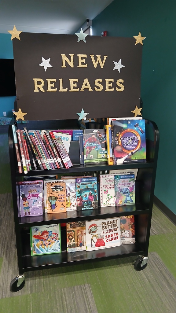 A black rolling bookshelf featuring a large black sign with gold glitter letters that reads "NEW RELEASES" adorned with gold, silver, and blue paper stars. The shelves are stocked with graphic novels and popular youth books, including Minecraft, Space, and sports titles.