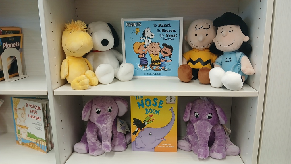 A white library bookshelf featuring plush toys and children's books. The top shelf displays a "Be Kind, Be Brave, Be You!" Peanuts book surrounded by plush Snoopy, Woodstock, Charlie Brown, and Lucy dolls. The bottom shelf displays "The Nose Book" placed between two matching purple plush elephants.