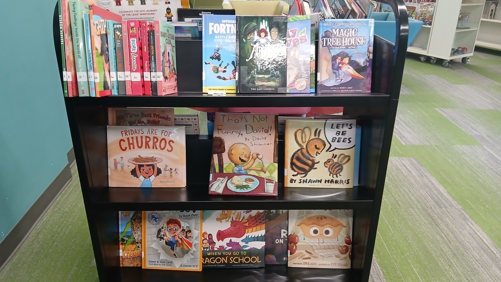 A black, three-tiered rolling bookshelf filled with children's books inside a brightly lit school library. Prominently displayed book covers include "Fridays are for Churros," "That's Not Funny, David!", and "Let's Be Bees."