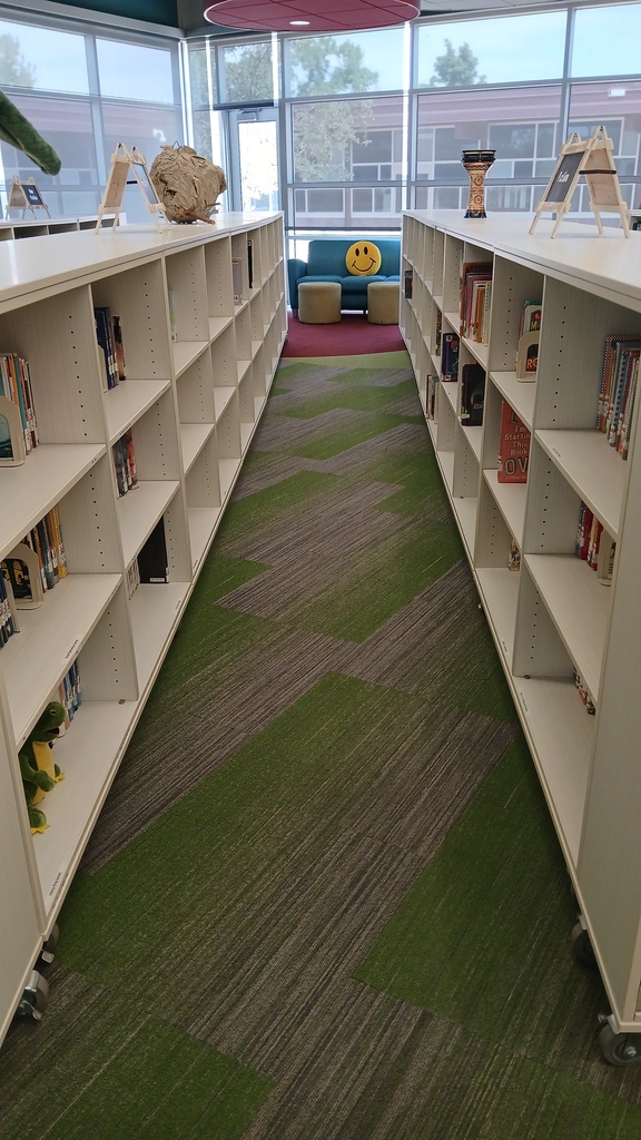 A view down a long, straight aisle formed by white, low-profile bookshelves in a modern library. The aisle features a patterned green and grey carpet leading toward a distant blue couch adorned with a bright yellow smiley-face pillow.