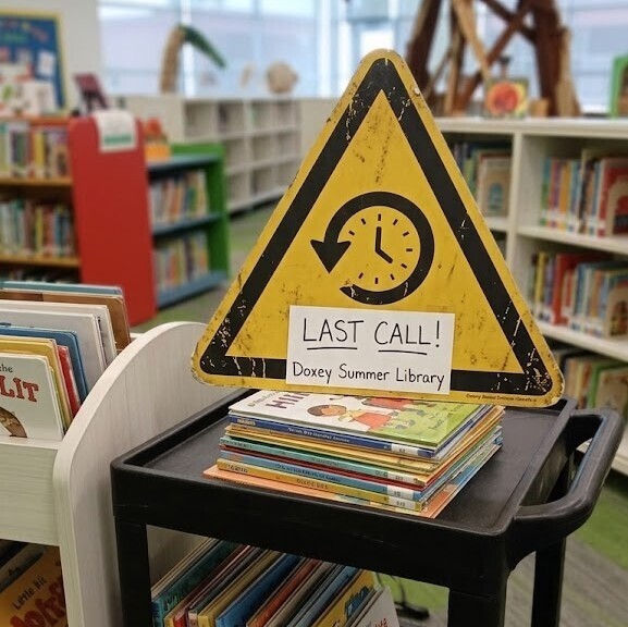 A yellow, triangular "LAST CALL!" caution sign with a clock icon is placed prominently on a stack of children's books sitting on top of a black utility cart. The sign text also includes "Doxey Summer Library" in smaller print. The scene is set inside the Doxey Elementary library, which is brightly lit and features rows of bookshelves and a distinctive wooden reading tree structure visible in the background