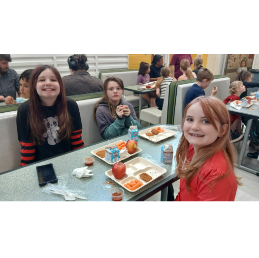 Three young girls smiling together at a cafeteria booth table set with lunch trays featuring red apples, baby carrots, milk, and chicken nuggets.