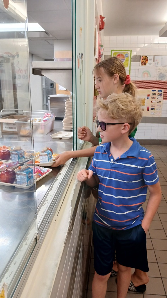 A boy wearing cool sunglasses and a blue striped polo shirt stands with a girl at the cafeteria service window, receiving a lunch tray packed with fresh apples, carrots, and milk cartons.
