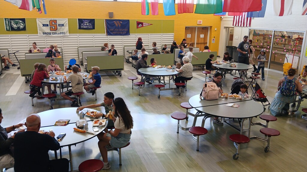 A wide, bright view of the Doxey Elementary cafeteria filled with children and adults sitting at round tables and booth seating, eating lunch together under decorative college flags hanging from the ceiling.