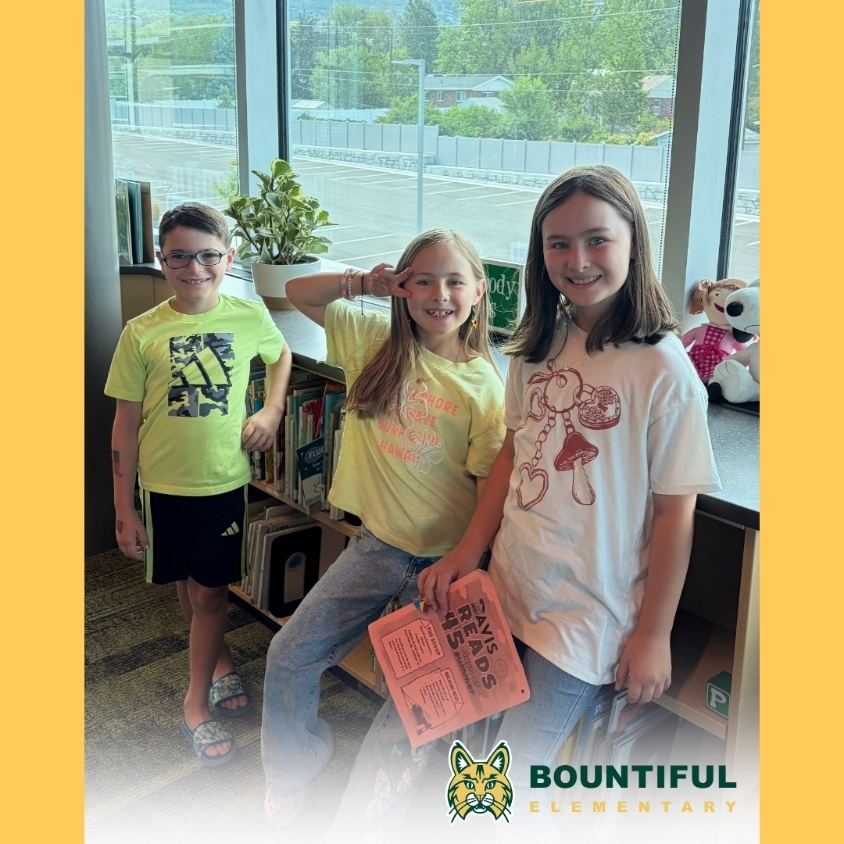 Three smiling Bountiful Elementary students stand together beside a bookshelf in the school library.