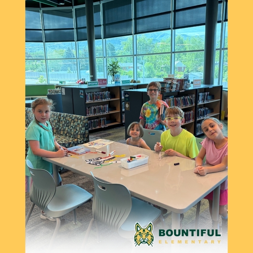Five students gather around a library table for a craft activity, with bookshelves and mountain views behind them.