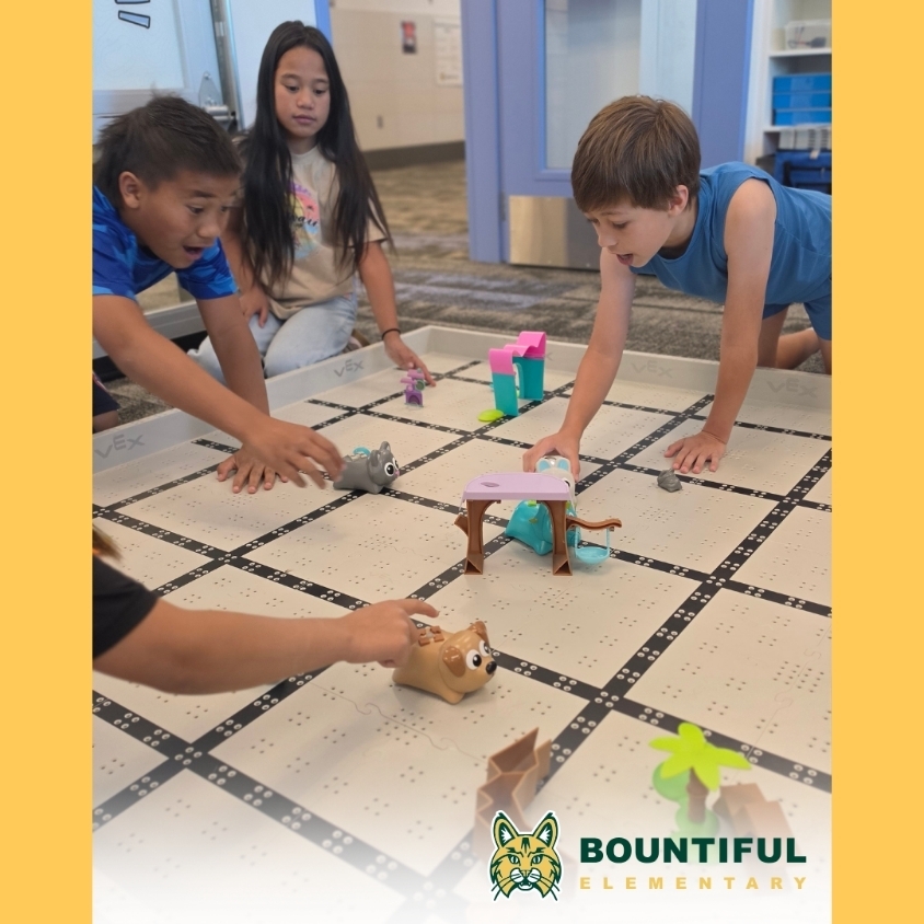 Students gather around a VEX field in the computer lab, using Coding Critters and small obstacles during a robot game activity.