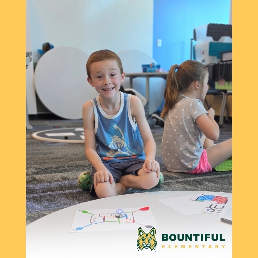 A student smiles beside an Ozobot path drawing while another student works nearby during a robot game activity.